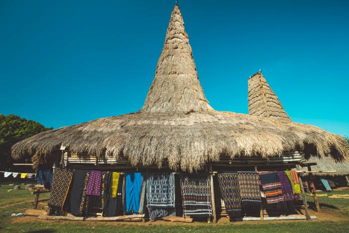 Traditional Sumba village house with tall thatched roof and ikat fabrics hanging outside in East Sumba.